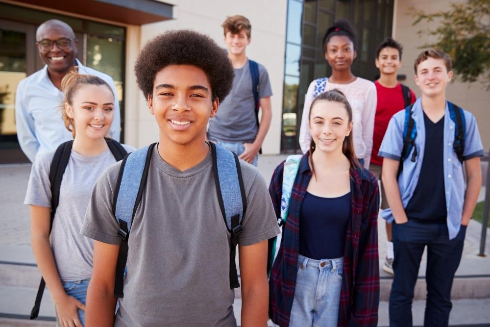 grupo de estudantes diversos sorrindo em frente à escola, acompanhados por um professor ao fundo.