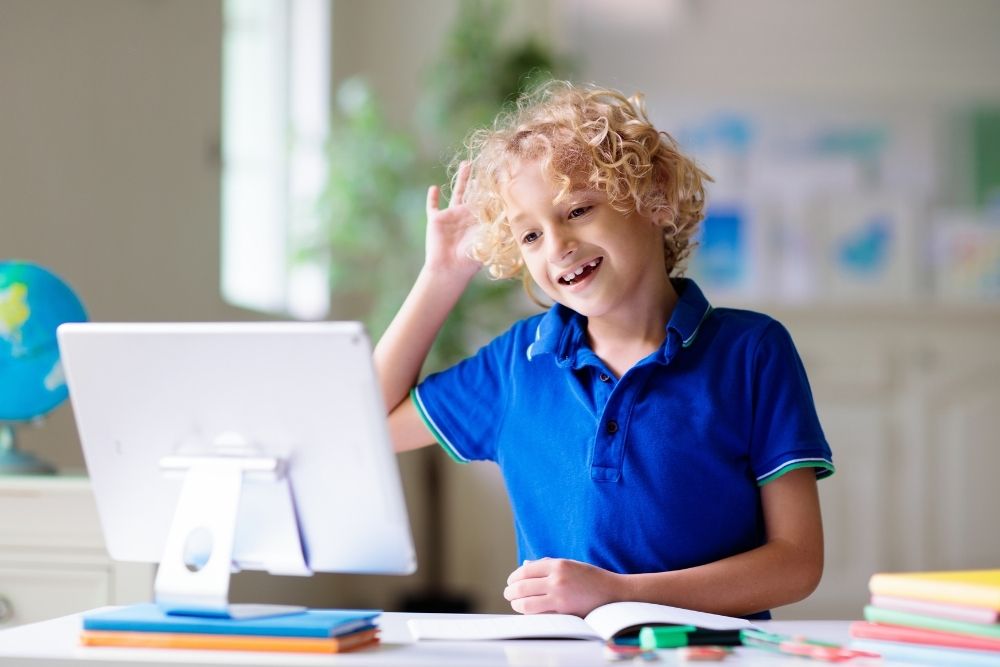 Criança estudando online em casa com computador e livros, aprendendo de forma interativa com apoio da tecnologia educacional.