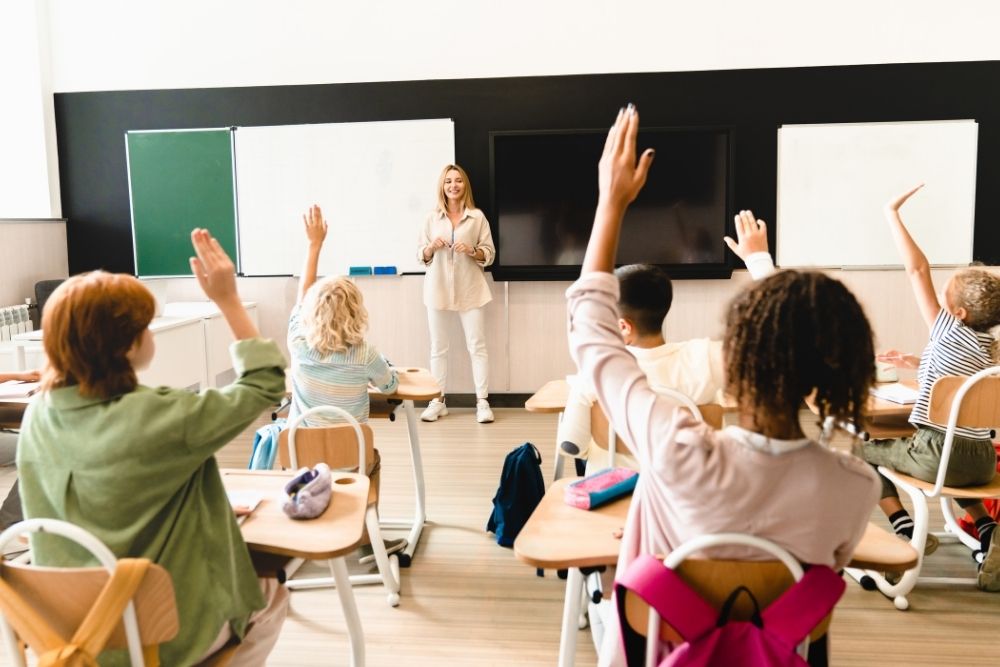 Uma sala de aula moderna e bem iluminada com uma professora sorridente, em pé na frente de um quadro negro. Ela está olhando para um grupo de alunos, que estão sentados em suas carteiras, com a maioria deles com as mãos levantadas para fazer perguntas ou responder a uma questão.
