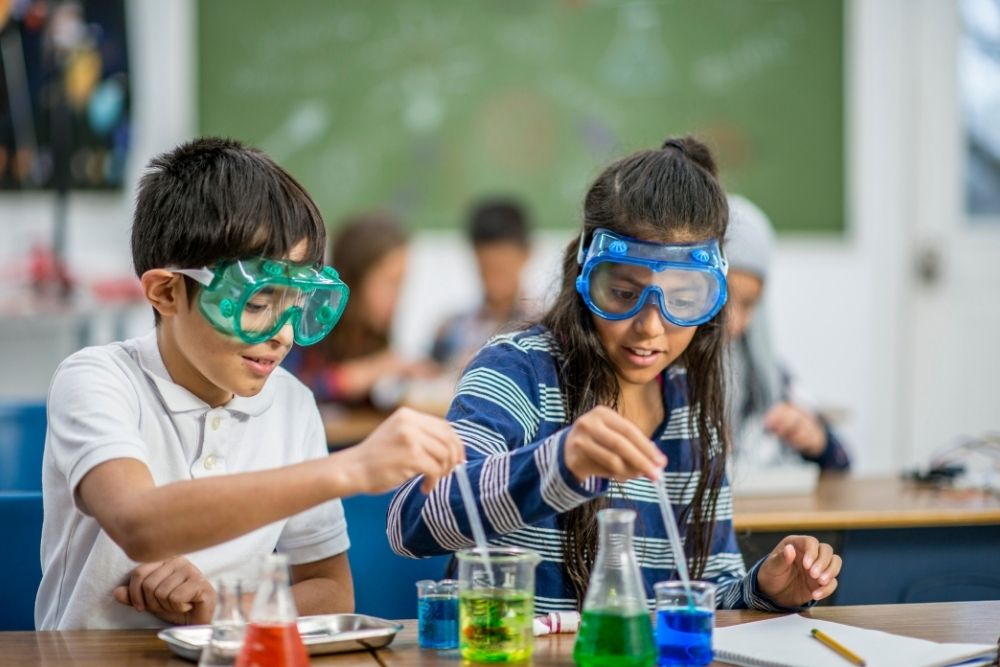 Dois estudantes, um menino e uma menina, usam óculos de proteção e trabalham em um experimento de ciências com tubos de ensaio e líquidos coloridos. Eles representam o lado acadêmico das olimpíadas estudantis.