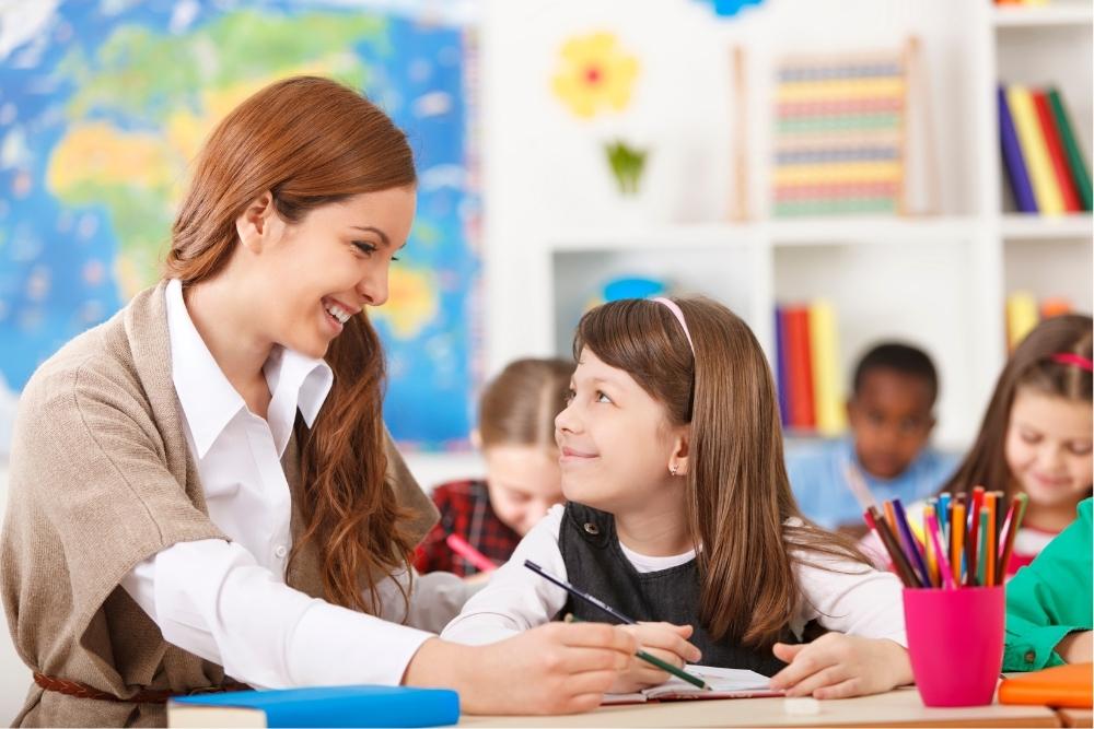 Uma professora sorridente e atenciosa se inclina para ajudar uma jovem aluna em sua carteira, estabelecendo contato visual e demonstrando a personalização do ensino.